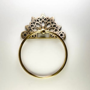 May include: A gold-toned towel ring with a decorative, ornate design. The ring is circular and has a detailed, intricate pattern.