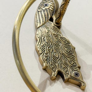 May include: A brass peacock-themed towel ring. The peacock is intricately detailed with etched feathers and a curved tail. The ring is a circular shape, and the fixture is mounted with screws.