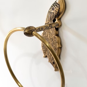 May include: A brass peacock-shaped towel ring. The peacock is detailed with etched feathers and a curved ring for holding a towel. The fixture is mounted on a white wall.