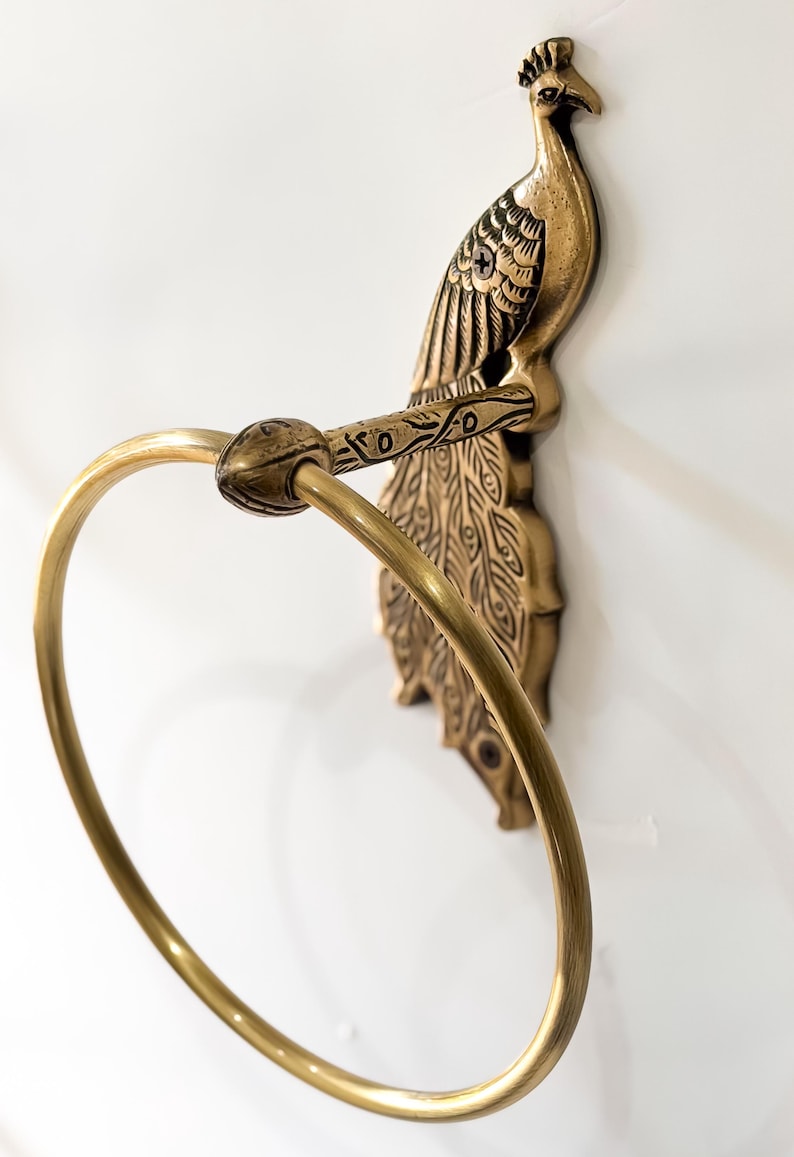 May include: A brass peacock-shaped towel ring. The peacock is detailed with etched feathers and a curved ring for holding a towel. The fixture is mounted on a white wall.