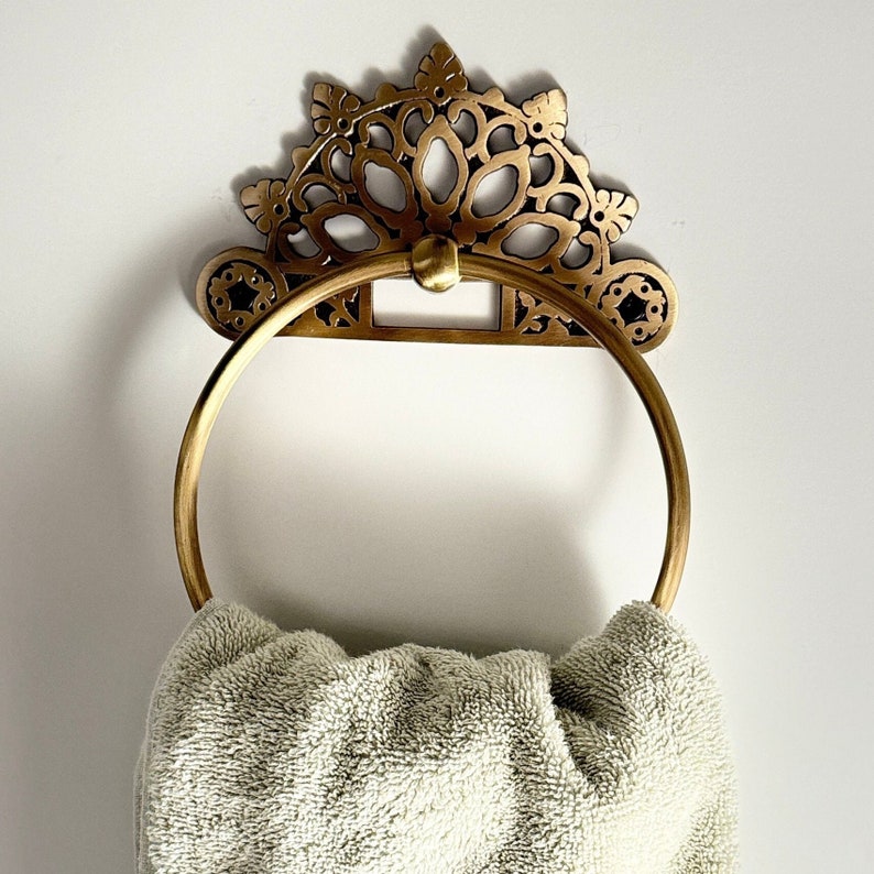 May include: A gold-toned towel ring with a decorative, ornate design. The ring is attached to a white wall and has a towel draped over it.
