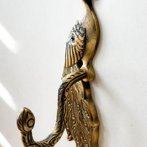 May include: A decorative brass peacock wall hook. The hook is shaped like a peacock with detailed feather and body patterns. The hook is mounted on a white wall.