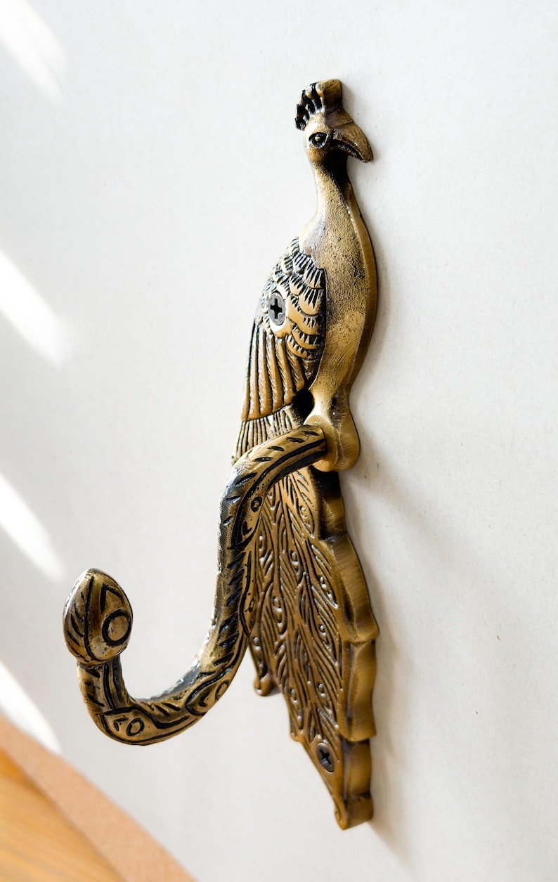 May include: A decorative brass peacock wall hook. The hook is shaped like a peacock with detailed feather and body patterns. The hook is mounted on a white wall.