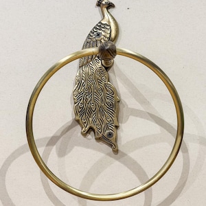 May include: A brass peacock-shaped towel ring. The peacock has detailed feather patterns and a circular ring for hanging towels. The fixture is mounted on a wall, showcasing a decorative and functional bathroom accessory.