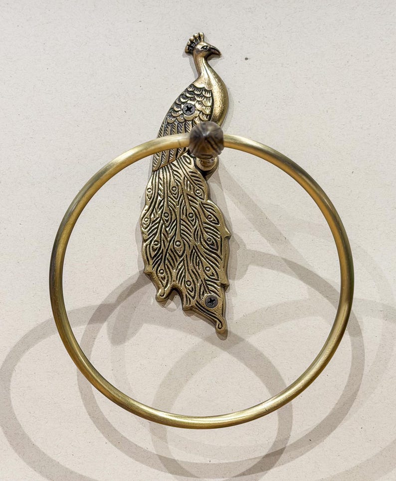 May include: A brass peacock-shaped towel ring. The peacock has detailed feather patterns and a circular ring for hanging towels. The fixture is mounted on a wall, showcasing a decorative and functional bathroom accessory.