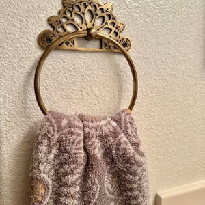 May include: A gold-toned metal towel ring with an ornate design hangs on a white wall. A brown towel is draped over the ring.