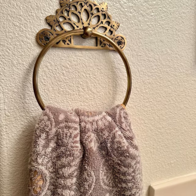 May include: A gold-toned metal towel ring with an ornate design hangs on a white wall. A brown towel is draped over the ring.