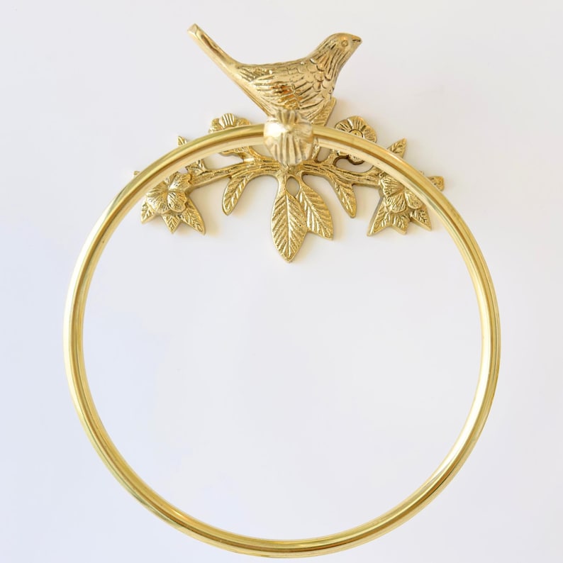 May include: A gold-colored towel ring with a bird perched atop a branch with leaves and flowers. The ring is circular and made of metal. The bird and branch are also made of metal and are attached to the wall.