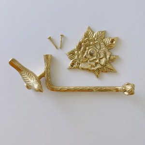May include: A gold-colored, decorative toilet paper holder set. The set includes a bird-shaped arm, a floral backplate, and two screws. The holder is made of metal and has a detailed, textured design.