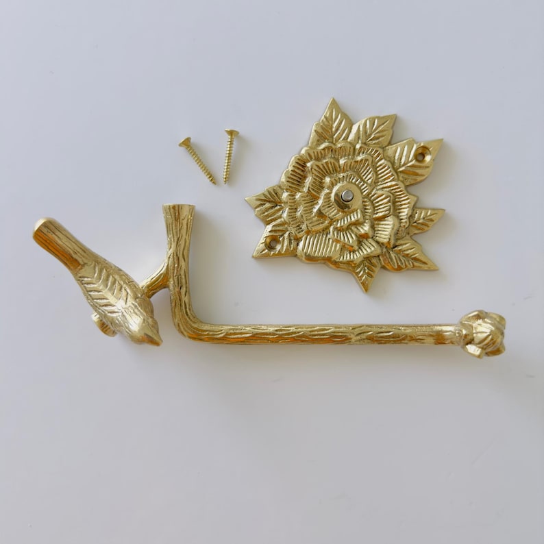 May include: A gold-colored, decorative toilet paper holder set. The set includes a bird-shaped arm, a floral backplate, and two screws. The holder is made of metal and has a detailed, textured design.