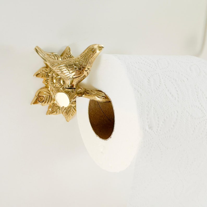 May include: A gold-colored metal toilet paper holder featuring a detailed bird perched on a branch with leaves. The holder is mounted on a white wall, holding a roll of white toilet paper. The bird and branch design adds a decorative touch to the bathroom.