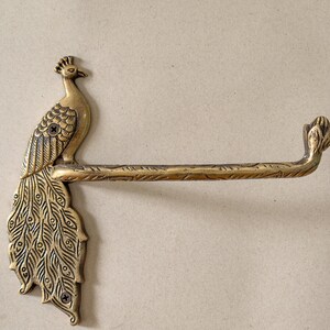 May include: A brass peacock toilet paper holder. The holder features a detailed peacock design with a long bar for holding the toilet paper roll. The peacock is perched on the left side of the holder, with its tail feathers fanned out.