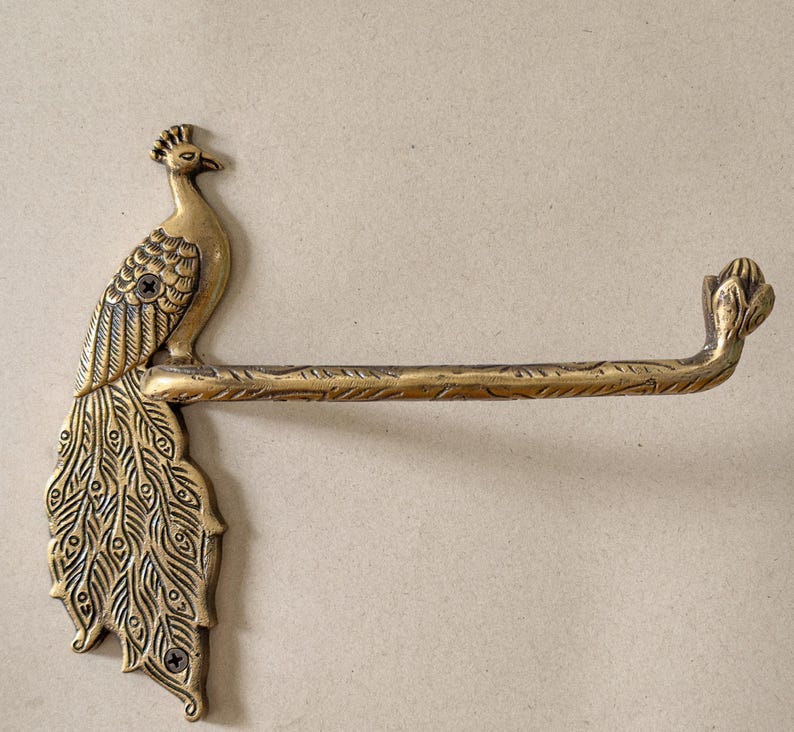 May include: A brass peacock toilet paper holder. The holder features a detailed peacock design with a long bar for holding the toilet paper roll. The peacock is perched on the left side of the holder, with its tail feathers fanned out.