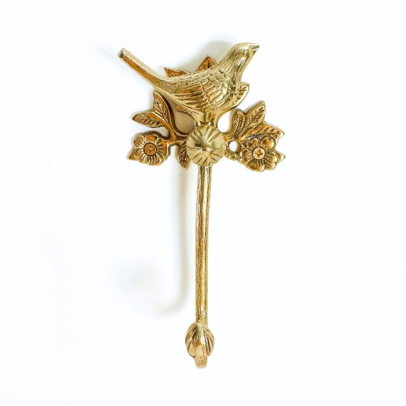 May include: A brass wall hook featuring a detailed bird perched on a floral branch. The hook has a long, slender stem ending in a decorative bud. The bird and floral details are in a gold finish.
