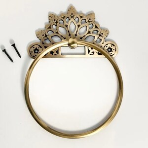 May include: Gold-toned metal towel ring with an ornate, decorative design. The ring is attached to a wall with two screws.