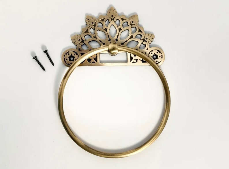May include: Gold-toned metal towel ring with an ornate, decorative design. The ring is attached to a wall with two screws.