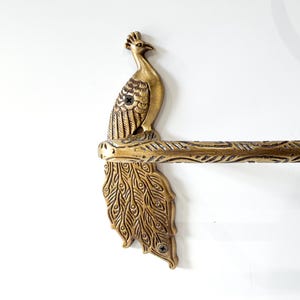 May include: A brass-colored peacock-themed towel rack. The peacock is perched atop a horizontal bar with a detailed feather design. The peacock's tail feathers are fanned out below the bar, adding to the decorative appeal. The rack is mounted on a white wall.