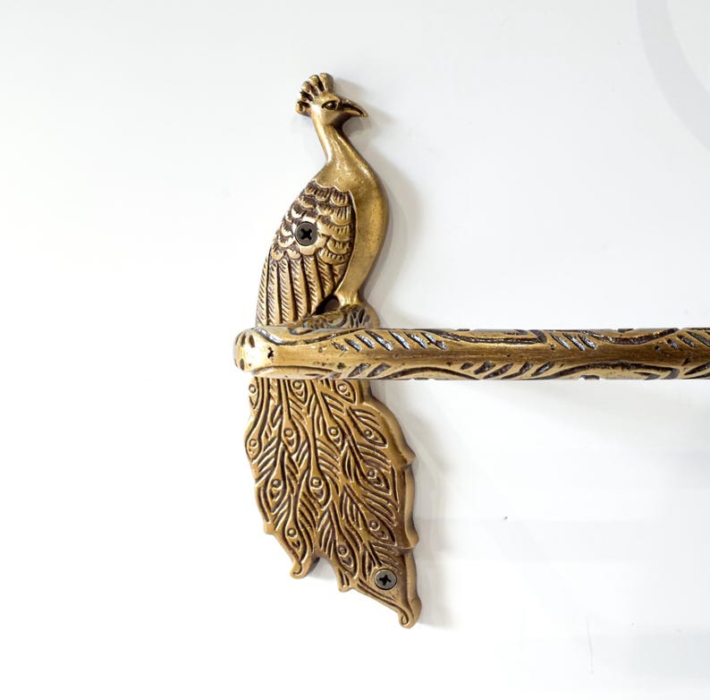 May include: A brass-colored peacock-themed towel rack. The peacock is perched atop a horizontal bar with a detailed feather design. The peacock's tail feathers are fanned out below the bar, adding to the decorative appeal. The rack is mounted on a white wall.