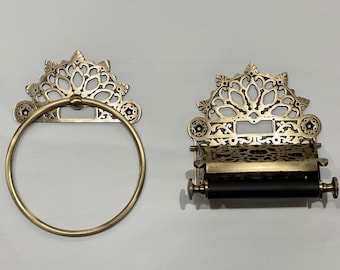 Op de afbeelding: Een handdoekring en een bijpassende toiletrolhouder van messingkleurig metaal. Beide items hebben een sierlijke, opengewerkte filigraan achterplaat met een decoratieve boogvormige bovenkant. De handdoekring is rond en de rolhouder heeft een stang.