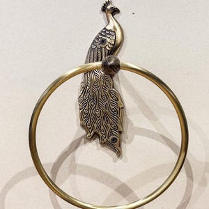 May include: A brass peacock towel ring. The peacock is detailed with engraved feathers and a crown. The circular ring is attached to the peacock's body. The metal has a vintage, antique finish. The towel ring is mounted on a light beige wall.