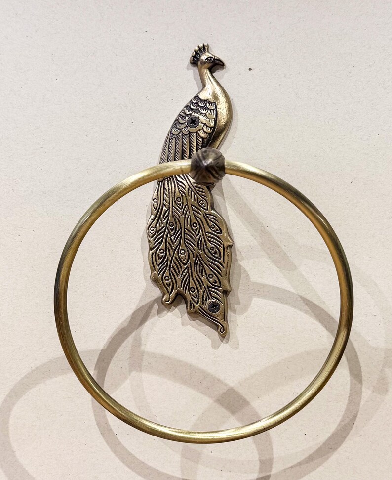 May include: A brass peacock towel ring. The peacock is detailed with engraved feathers and a crown. The circular ring is attached to the peacock's body. The metal has a vintage, antique finish. The towel ring is mounted on a light beige wall.