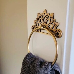 May include: Gold-toned decorative towel ring with an ornate, floral design. A gray towel is hanging on the ring.