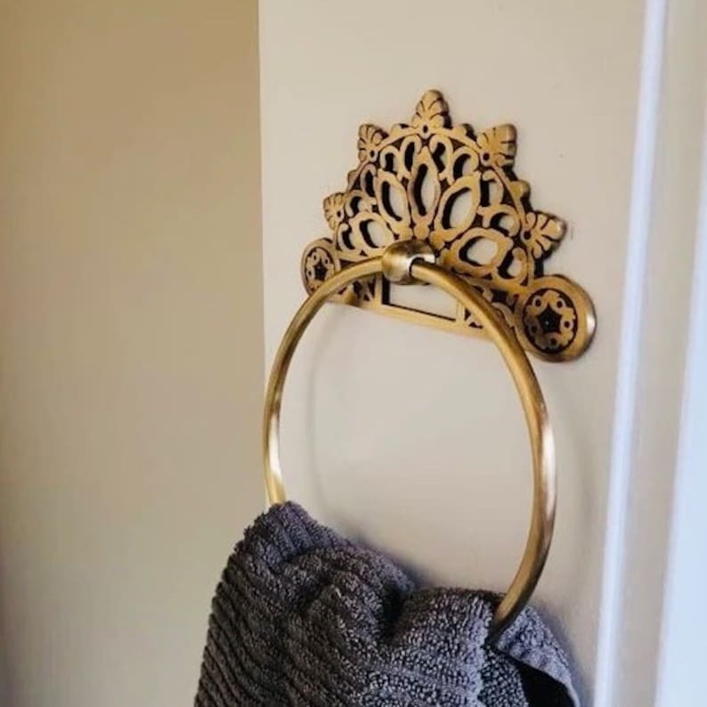 May include: Gold-toned decorative towel ring with an ornate, floral design. A gray towel is hanging on the ring.