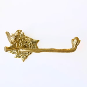 May include: A brass-colored toilet paper holder featuring a bird perched on a branch. The holder has a floral and leaf design, with a branch-shaped arm extending to hold the toilet paper roll. The overall design is ornate and decorative.