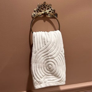 May include: A white towel with a swirling pattern hangs on a gold ornate towel ring.