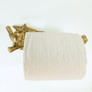 May include: A gold-colored, bird-shaped toilet paper holder. The holder is mounted on a white wall and holds a roll of white toilet paper. The bird is perched on a branch with leaves.