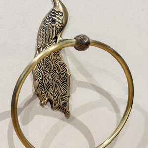 May include: A brass peacock towel ring. The peacock is detailed with etched feathers and a decorative head. The ring is attached to the peacock's body and is designed to hold a towel. The fixture is mounted on a neutral-colored wall.