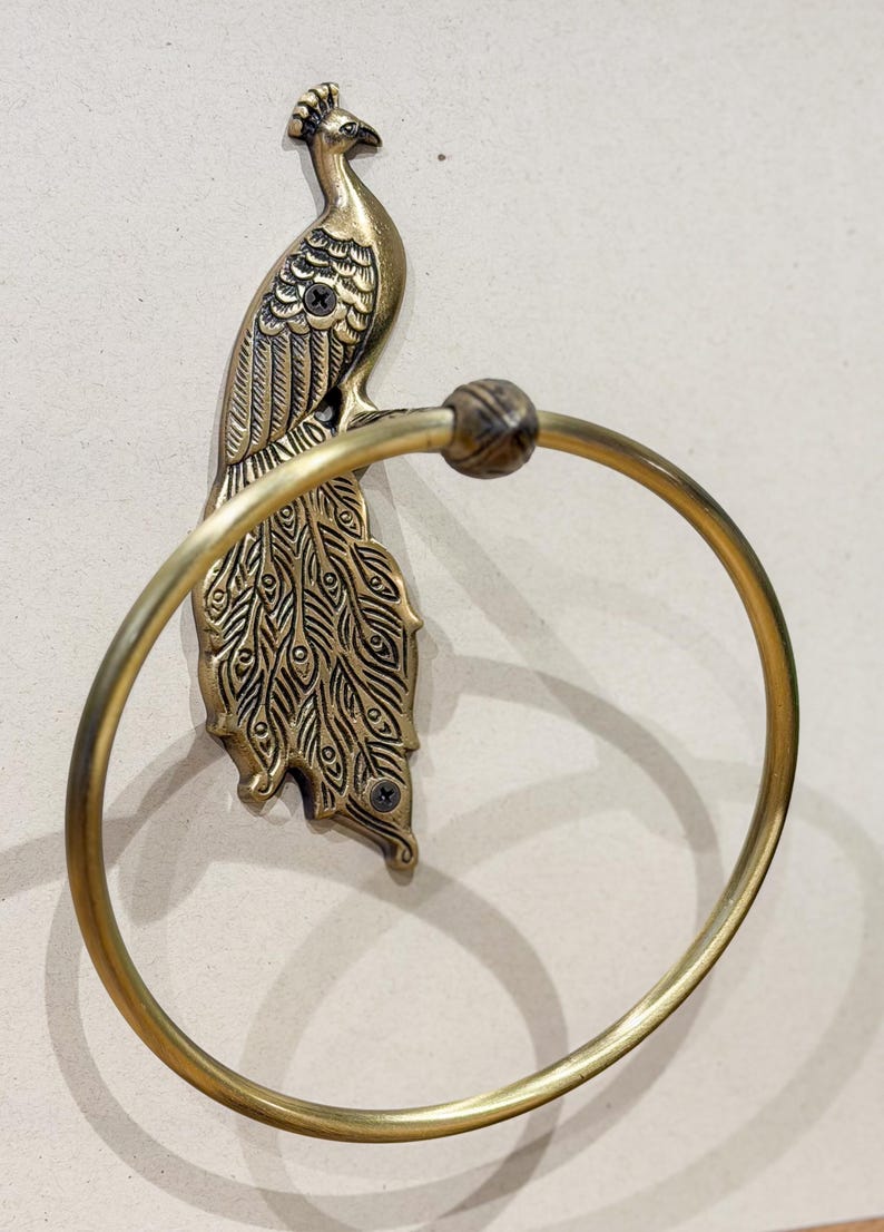 May include: A brass peacock towel ring. The peacock is detailed with etched feathers and a decorative head. The ring is attached to the peacock's body and is designed to hold a towel. The fixture is mounted on a neutral-colored wall.