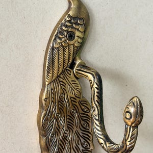 May include: A brass peacock wall hook. The hook is shaped like a peacock with detailed feather and body designs. The hook extends from the peacock's body in a curved shape. The hook is mounted with a screw.
