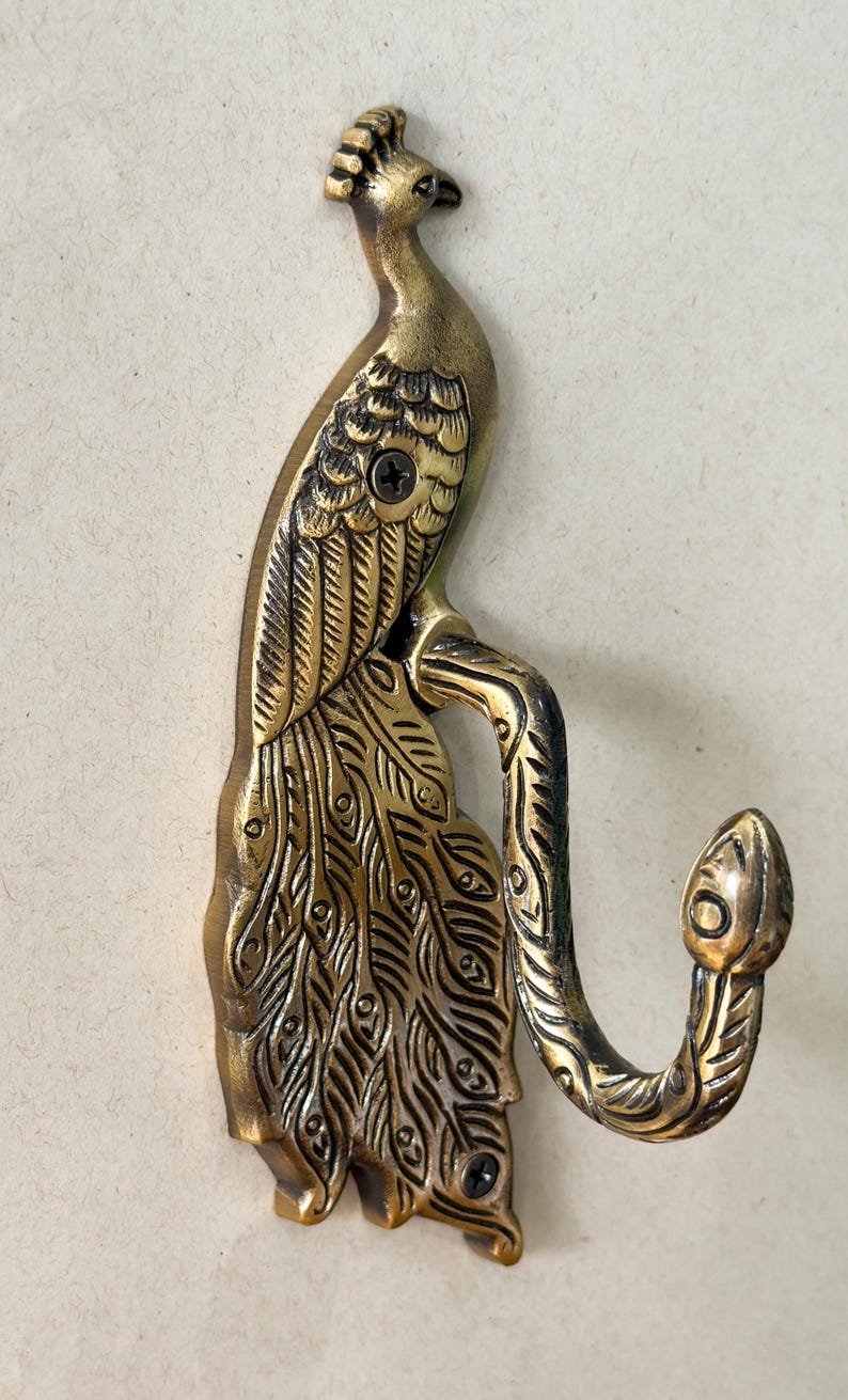 May include: A brass peacock wall hook. The hook is shaped like a peacock with detailed feather and body designs. The hook extends from the peacock's body in a curved shape. The hook is mounted with a screw.