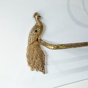 May include: A brass towel bar featuring a decorative peacock-shaped mounting bracket. The peacock has detailed feather engravings and is attached to a textured horizontal bar. Two visible screws secure the peacock bracket to the wall.