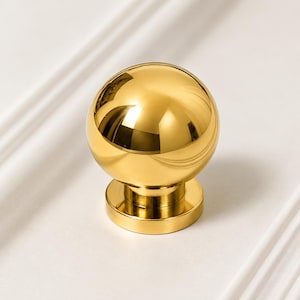 Könnte beinhalten: Ein polierter goldener Möbelknopf mit einem kugelförmigen Kopf auf einem zylindrischen Sockel. Die metallische Oberfläche hat ein reflektierendes Hochglanz-Finish, das das Licht einfängt und helle Glanzlichter auf der runden Form erzeugt.