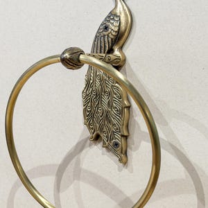 May include: A brass-colored towel ring featuring a peacock design. The peacock is perched atop a decorative backplate with a circular ring for hanging towels. The metal has a vintage finish, adding a touch of elegance to any bathroom.