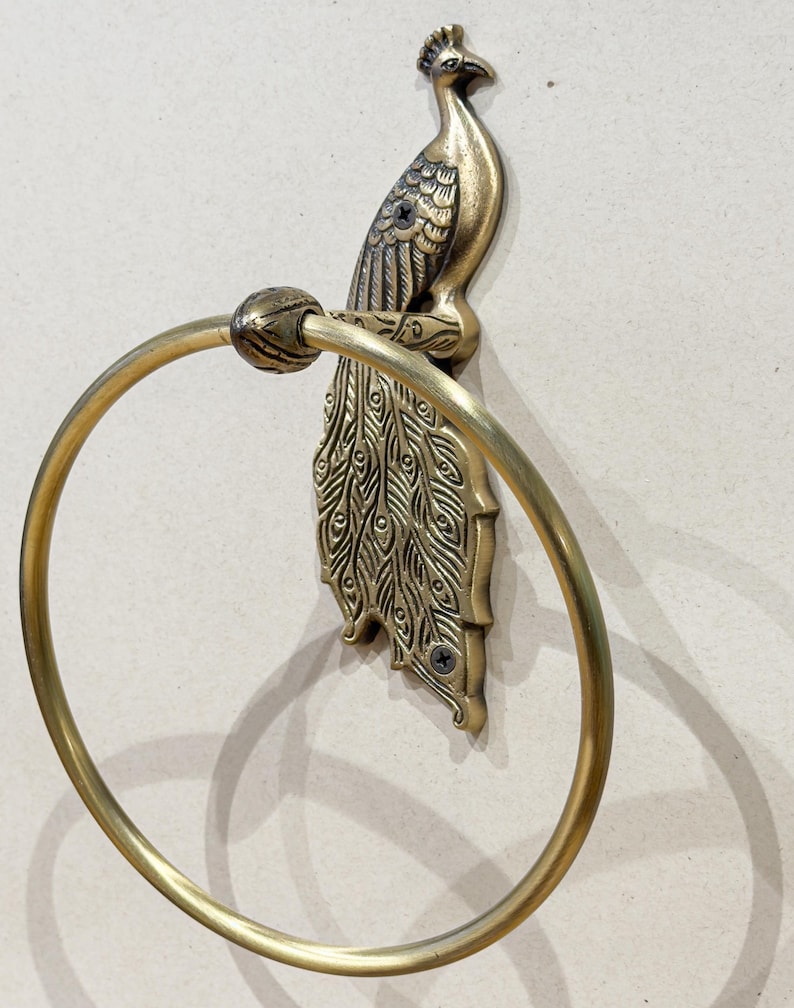 May include: A brass-colored towel ring featuring a peacock design. The peacock is perched atop a decorative backplate with a circular ring for hanging towels. The metal has a vintage finish, adding a touch of elegance to any bathroom.