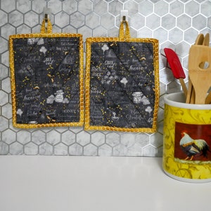 Puede incluir: Dos agarraderas de olla acolchadas negras y doradas con un tema de abejas y miel. Las agarraderas están colgadas en una pared de azulejos hexagonales grises. Un portautensilios de cerámica amarillo con un diseño de gallo está sobre una encimera blanca.