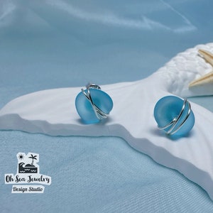 Può includere: Un paio di orecchini a bottone in vetro marino blu chiaro con avvolgimento in filo d'argento. Gli orecchini sono esposti su una superficie bianca a forma di onda, con una stella marina e il logo "Oh Sea Jewelry Design Studio".