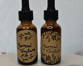 May include: Two amber glass bottles with black droppers. Each bottle has a tan label with the words "Yarrow Tincture" in cursive and a botanical illustration. The bottles are on a white surface.