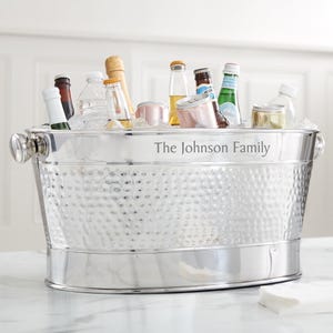 May include: A stainless steel beverage tub filled with ice and various bottles and cans. The tub has a hammered texture and the text "The Johnson Family" is printed on the side. The tub has two handles and is sitting on a white surface.