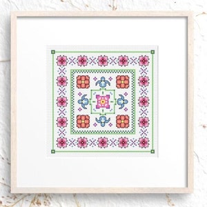 May include: A framed cross-stitch design featuring a square pattern with floral and geometric elements. The design includes pink and orange flowers, blue turtles, and a central square with a pink and yellow floral motif. The frame is light wood.