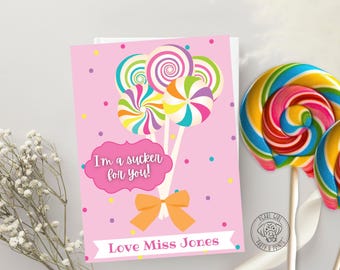 May include: A pink greeting card with a lollipop design and the text "I'm a sucker for you!" and "Love Miss Jones". Two large, colorful lollipops are on the right. White flowers are on the left.
