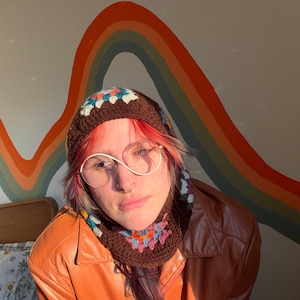 May include: A brown crochet hat with a colorful floral design. The hat is worn with a brown leather jacket and round, wire-rimmed glasses. A rainbow mural is in the background.