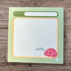 Peut inclure: Un bloc-notes vert avec un intérieur blanc à pois et une bordure jaune. Le bloc-notes a un en-tête vert avec un espace blanc vide. Le bas du bloc-notes a un cerveau rose avec un visage et le texte "NO MORE BRAIN FARTS".