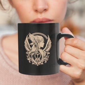 May include: Black ceramic mug featuring a detailed beige illustration of a woman with wings, a helmet, and ornate floral elements. The mug has a black handle and a glossy finish, suitable for hot beverages.