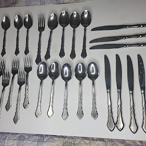 Könnte beinhalten: Eine Sammlung von Edelstahlbesteck, darunter Löffel, Gabeln und Messer, auf einer weißen Oberfläche angeordnet. Das Besteck hat ein dekoratives Griffdesign. Das Set umfasst verschiedene Größen von Löffeln, Gabeln und Messern, geeignet für ein formelles Esszimmer.