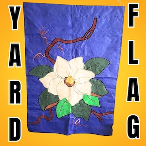 May include: A blue yard flag featuring a large white magnolia flower with green leaves and brown branches. The flag has the words "YARD FLAG" in large, bold, black letters on a yellow background.