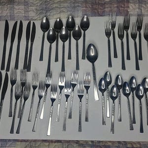 May include: A large collection of stainless steel flatware arranged on a white surface, including various dinner knives, dinner forks, salad forks, and spoons. The set features a mix of plain handles and pieces with decorative embossed patterns.